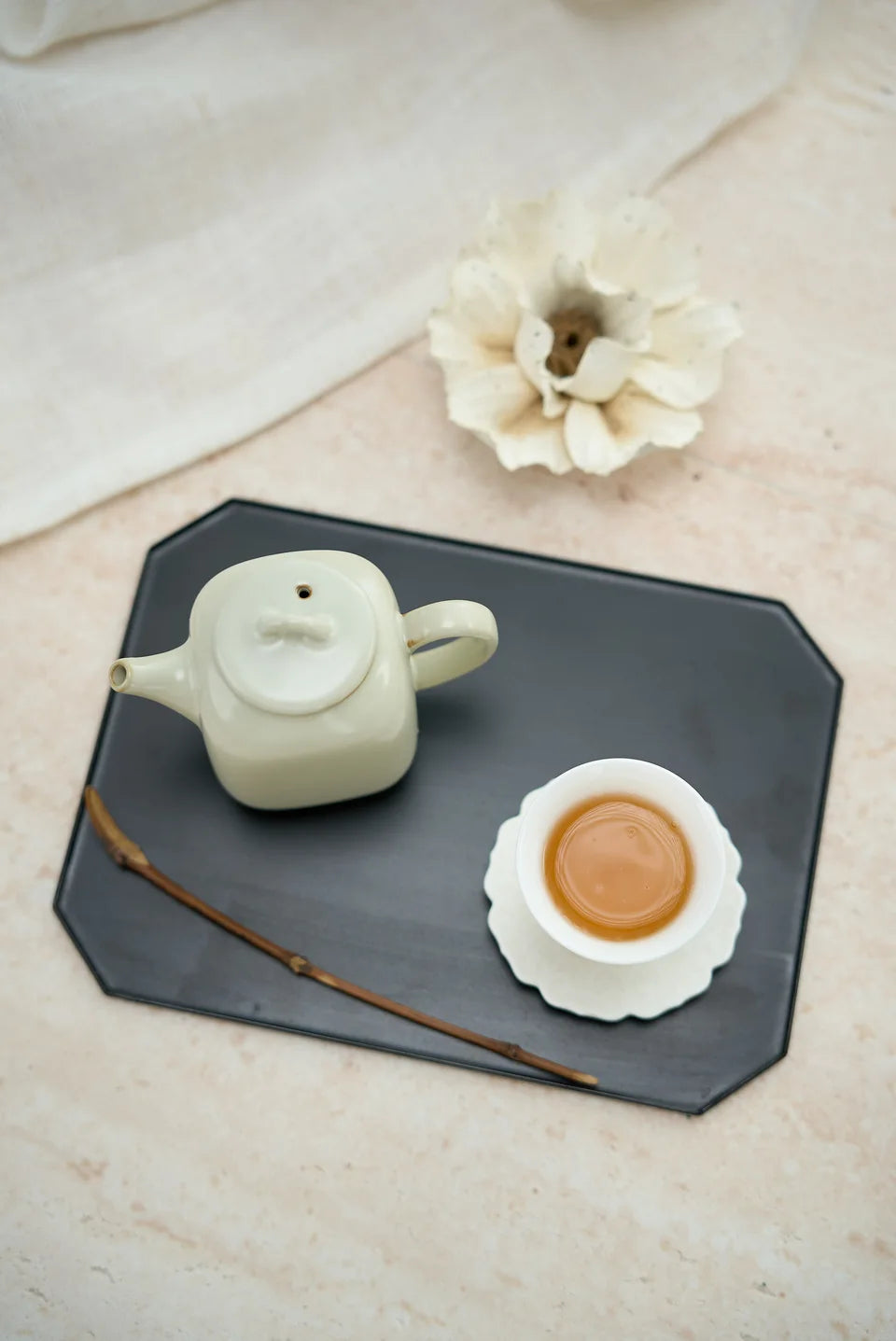 A pale teapot and a white cup of amber tea on a dark tray.