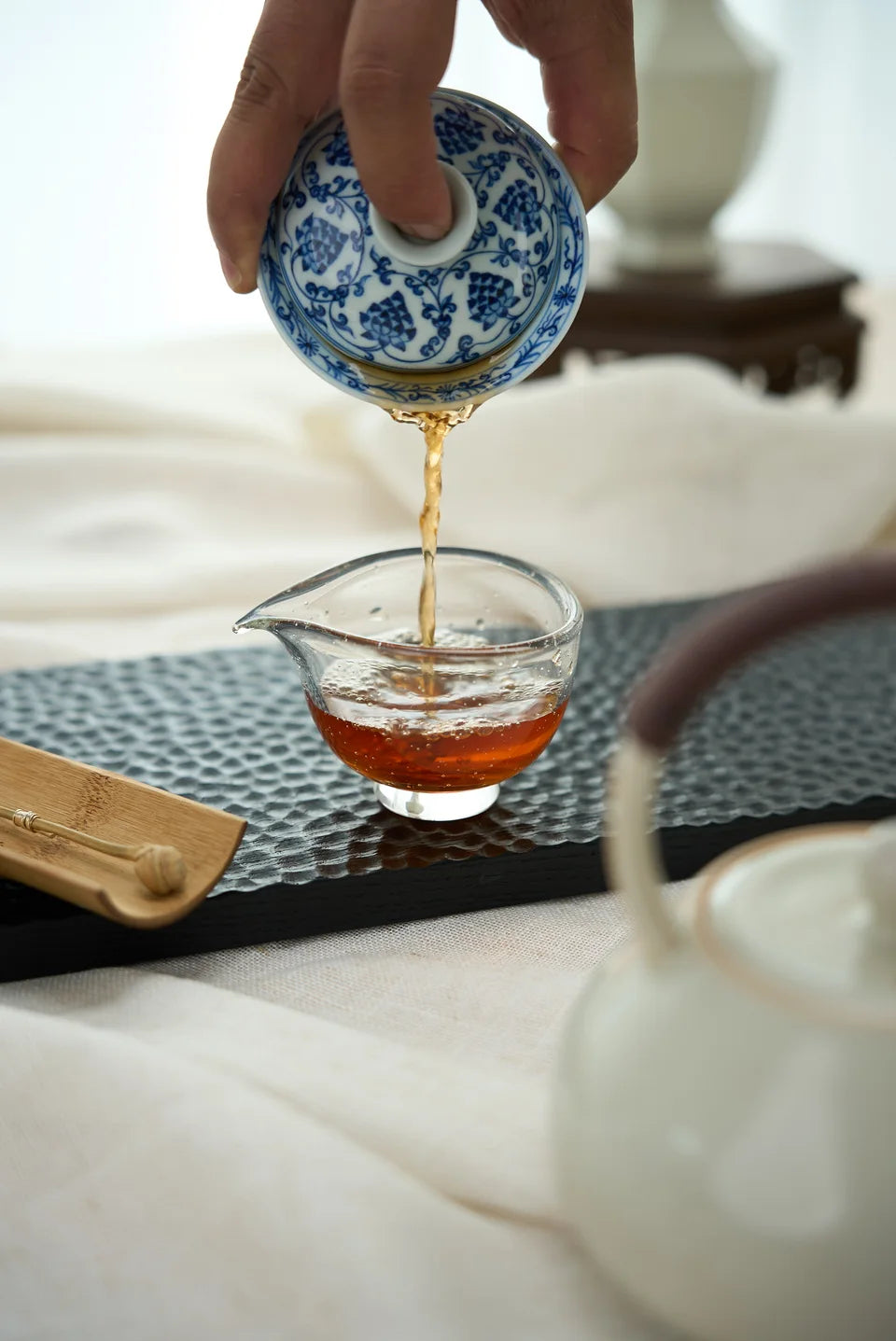 A hand pouring tea from a blue-and-white gaiwan into a glass pitcher.