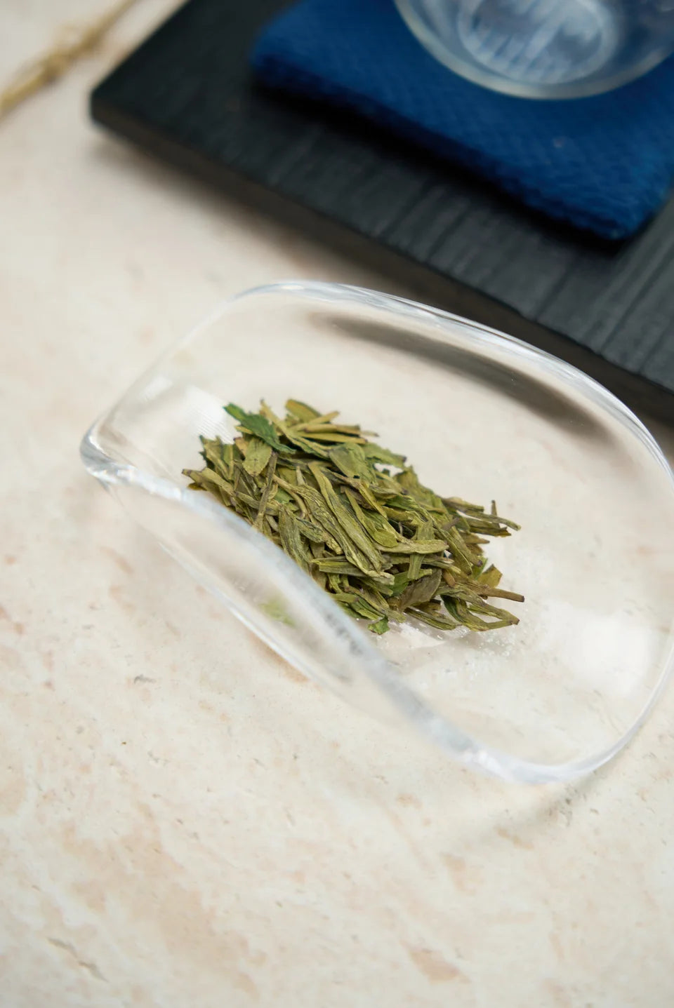 Flat green leaves in a glass tea scoop beside a blue tea cloth.