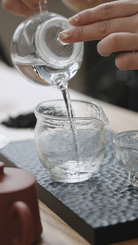 Clear water being poured from an empty glass teapot into a serving pitcher.