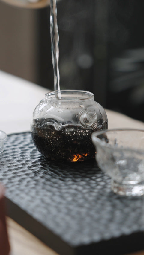 Water being poured into a small glass teapot containing tea leaves.