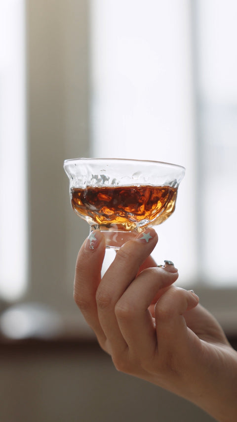 A hand holding a small glass cup of amber tea.