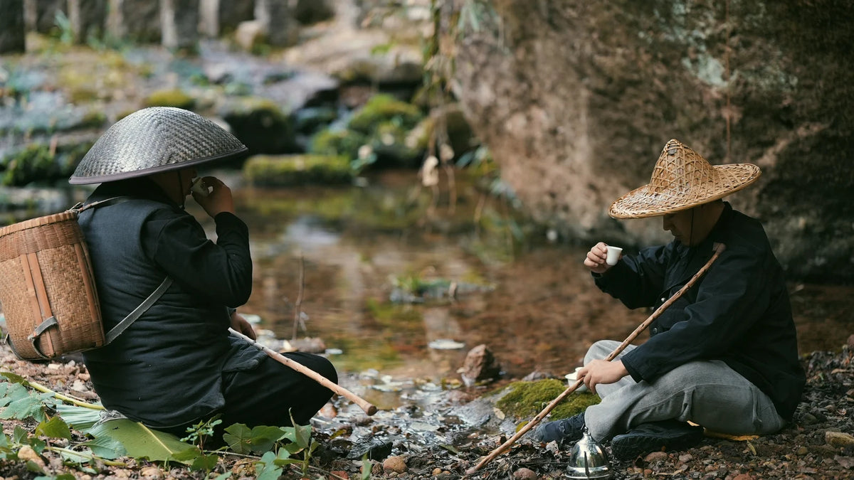 Two people sharing tea beside a stream beneath the trees