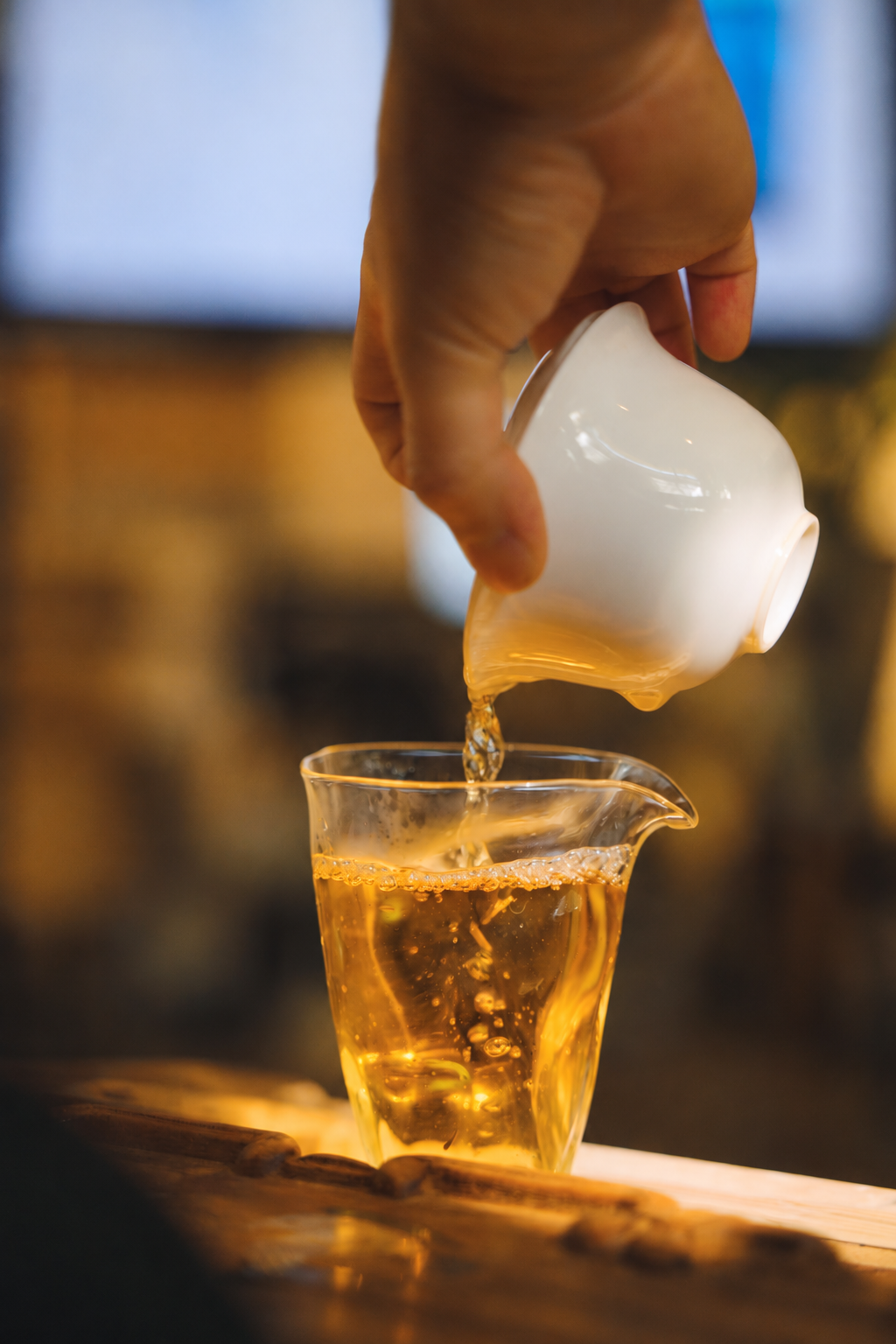 A hand pouring golden tea from a white gaiwan into a clear pitcher