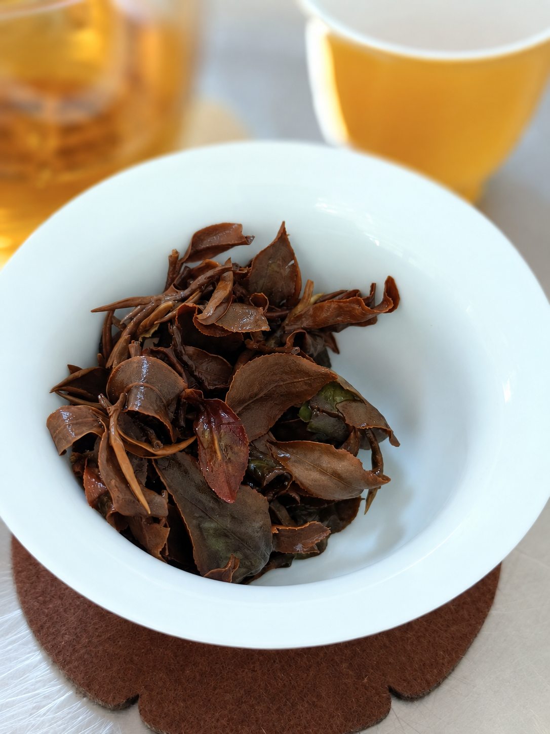 Opened brown tea leaves in a white bowl with amber tea behind