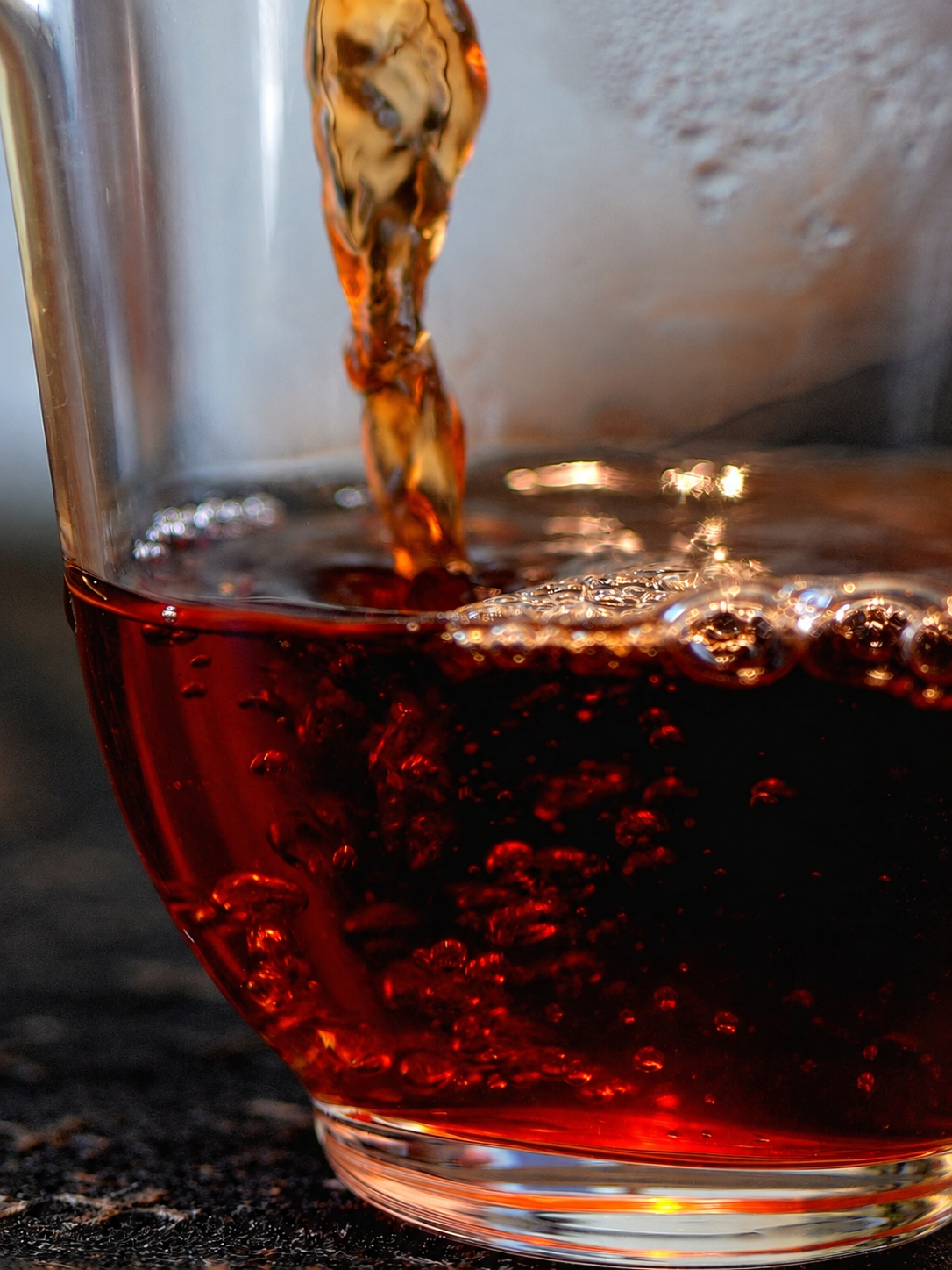 Reddish brown tea flowing into a clear glass vessel with bubbles at the surface