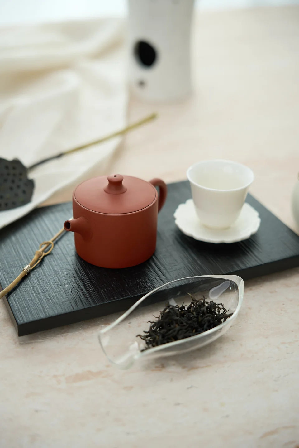 A red clay teapot and white cup beside dark loose tea in a glass scoop.