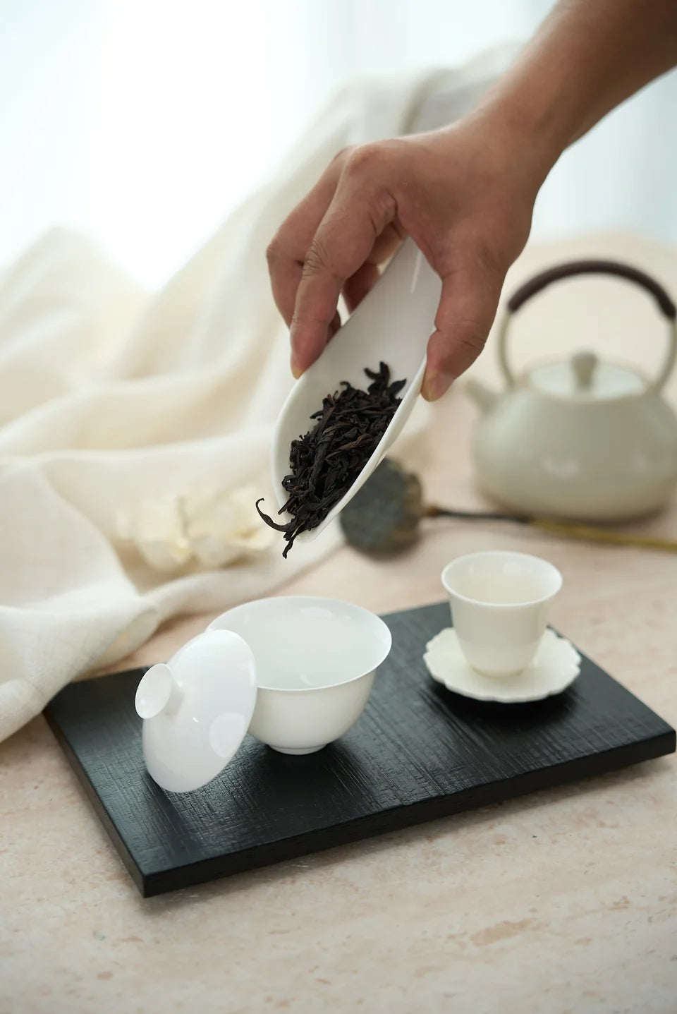 A hand tipping dry leaves from a white tea scoop into a white gaiwan.