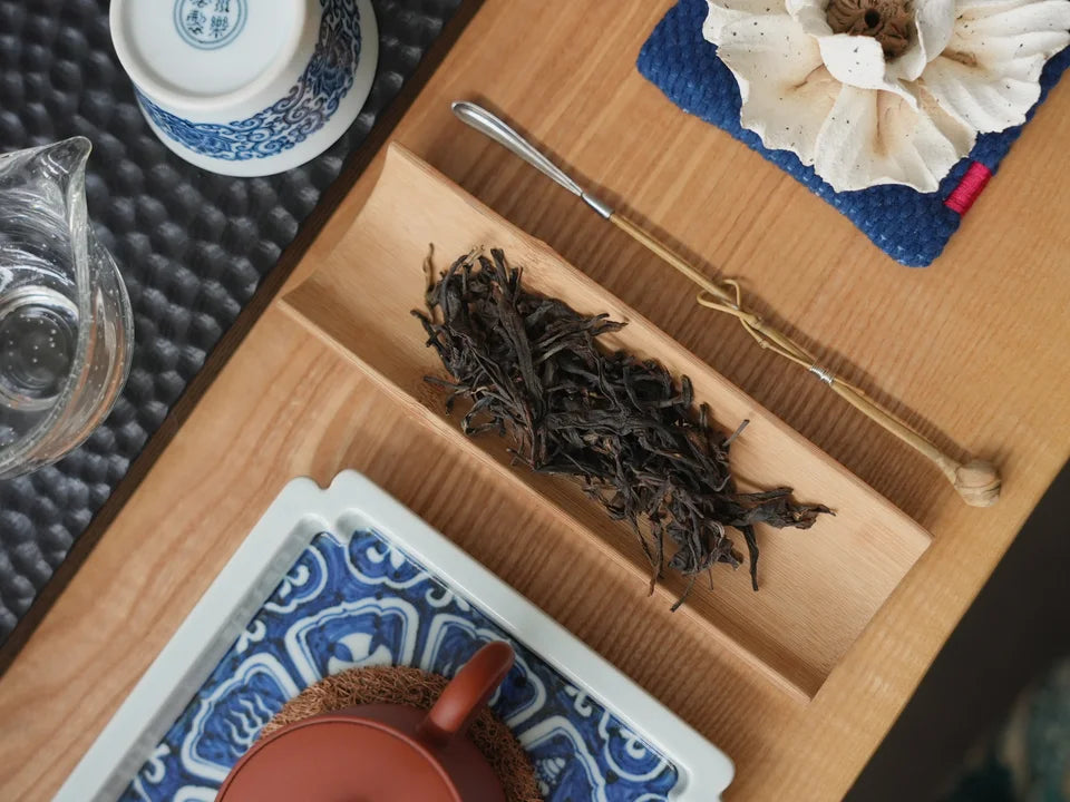 Loose raw Pu-erh leaves on a bamboo scoop beside a red clay teapot.
