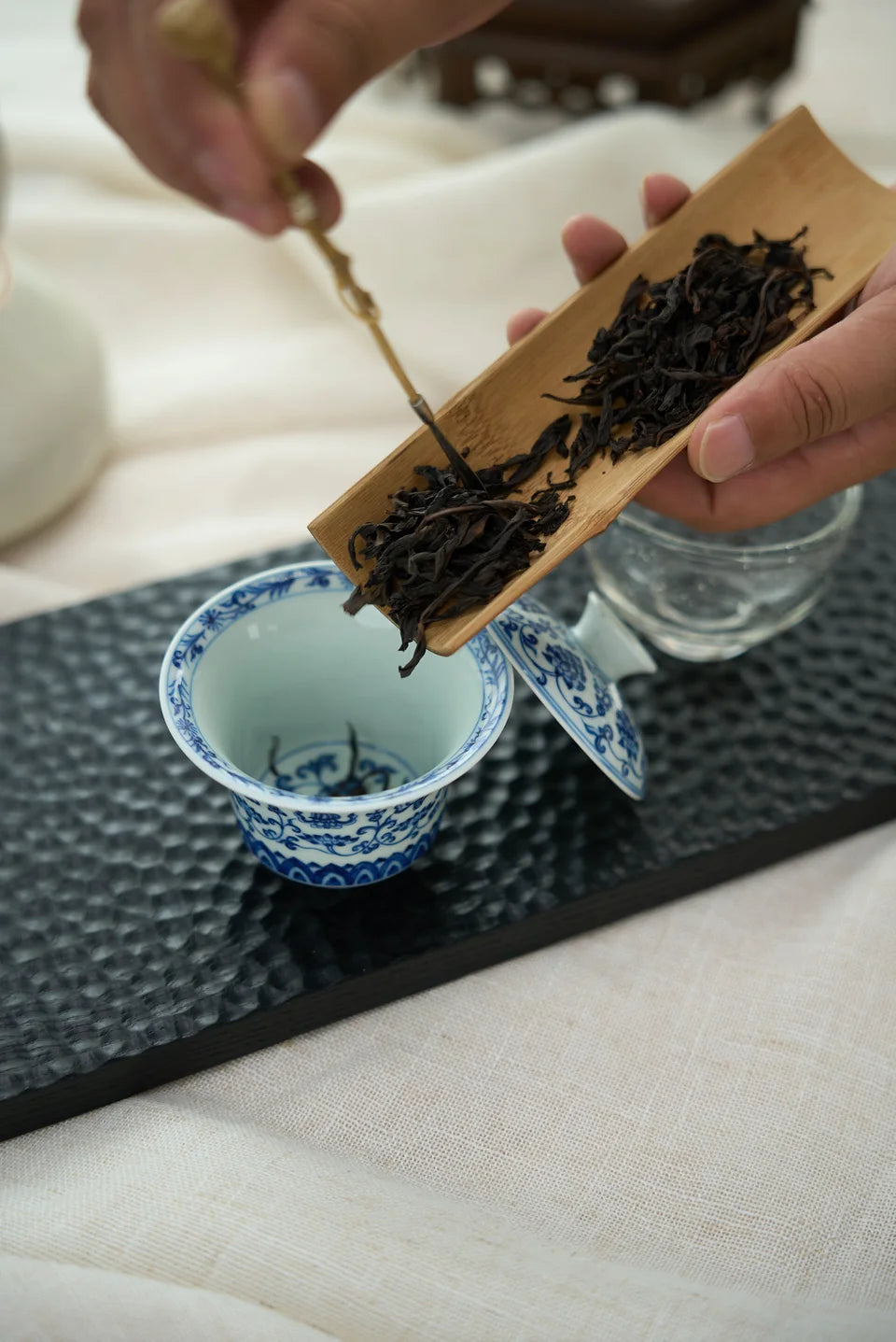 A hand guiding dry leaves from a scoop into a blue-and-white gaiwan.