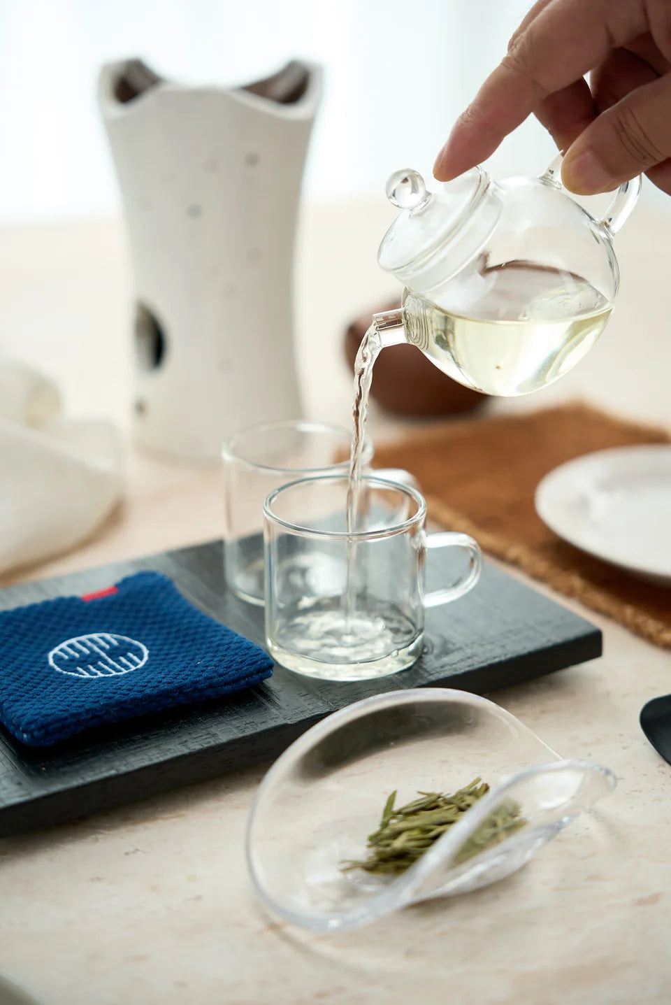 A hand pouring pale tea from a glass pitcher into a glass cup.