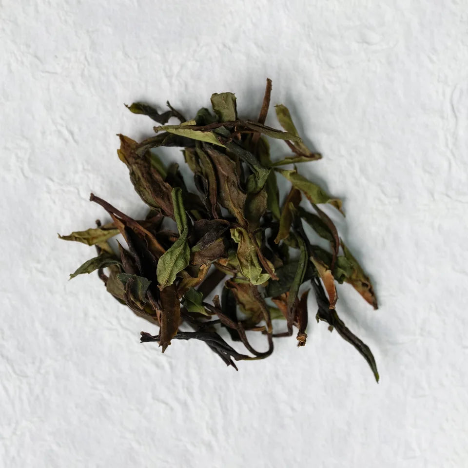 Loose white-tea leaves with mixed green and brown tones on a pale surface.