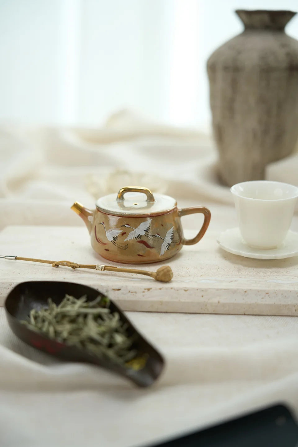 Pale tea buds in a dark scoop beside a decorated teapot and white cup.