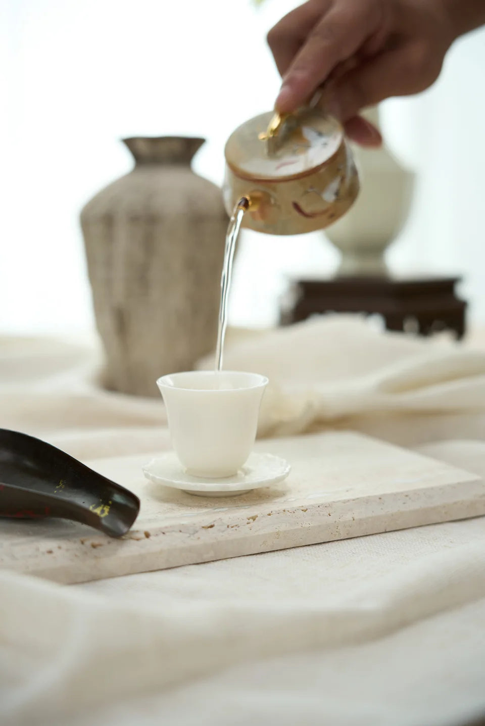A hand pouring from a decorated teapot into a small white cup.