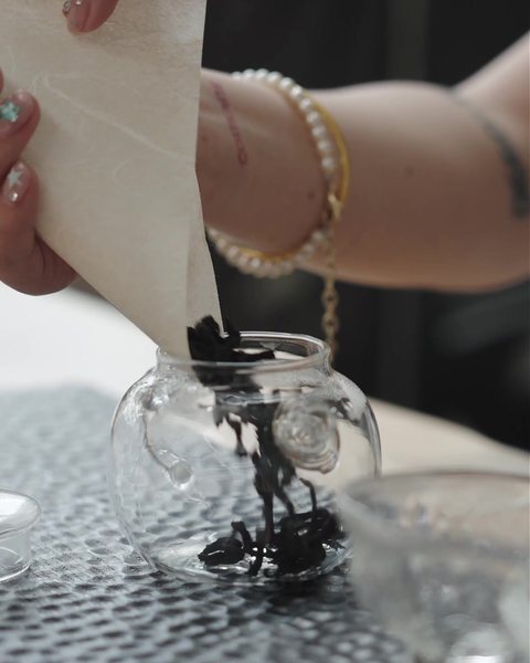 Loose tea leaves falling from folded paper into a glass teapot
